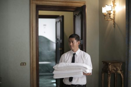 Housekeeper needed in Liverpool Young male housekeeper carrying stack of white bed sheets while entering bedroom in hotel
