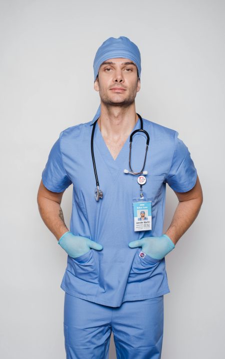 Registered Nurse needed in Wrexham (Visa Sponsorship Available) Male nurse in blue scrubs confidently posing with hands in pockets, stethoscope around neck, on white background.