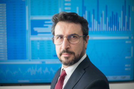 Professional man in suit and glasses stands confidently before stock market data screen.