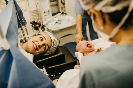 A mother smiles lovingly at her newborn in a hospital delivery room, attended by medical staff.