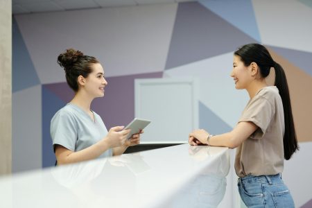 Admin/Receptionist needed in Manchester (Visa Sponsorship Available) A receptionist and a visitor engaging at a modern reception desk, indoors.