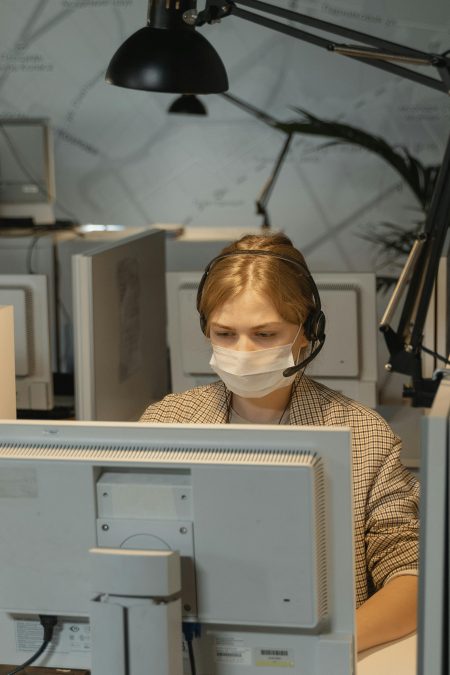 Call center employee wearing mask and headset, focused on computer work.