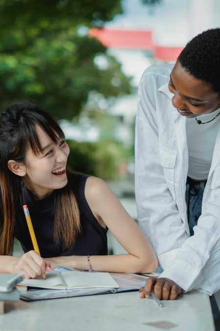 Happy young multiethnic ladies in casual outfit tutoring with books outside in summer day at table