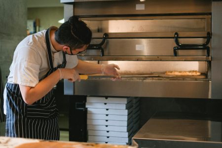 A chef in an apron prepares pizza in a commercial kitchen oven.