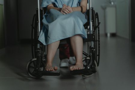 A person in a wheelchair, barefoot, wearing a hospital gown sitting in a dimly lit corridor.
