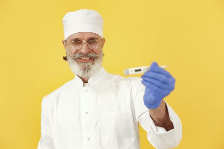 Elderly male doctor in white uniform with a digital thermometer and gloves against yellow background.