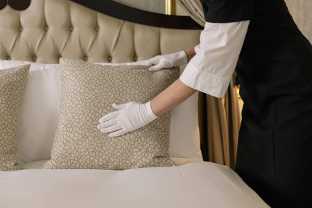 Housekeeping Assistant needed Dorset (Visa Sponsorship Available) A housekeeper wearing gloves arranges pillows on a luxurious bed in a hotel room.