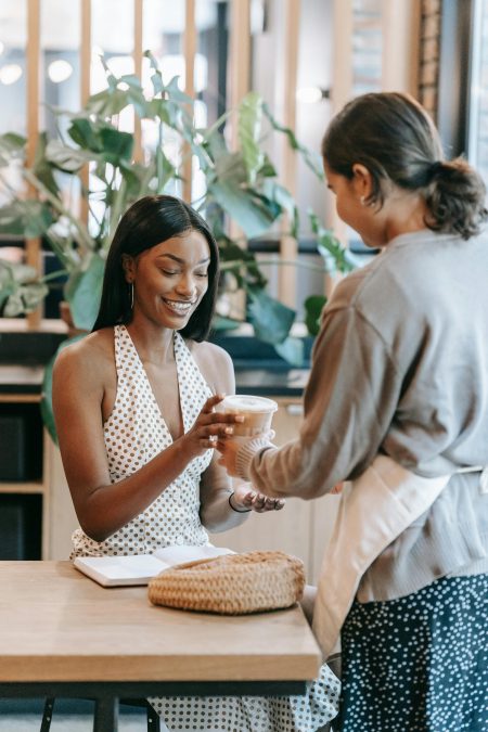 A woman in a polka-dot dress smiling as she receives a coffee from a barista in a stylish cafe.