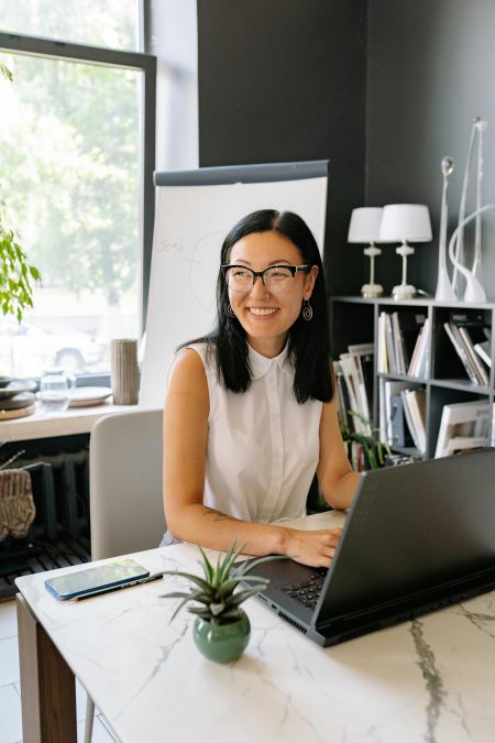 Smiling Asian businesswoman using laptop in a stylish, well-lit office with plants.