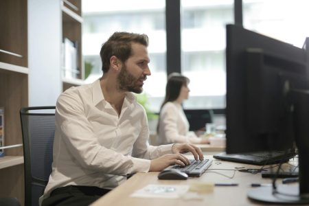 Side view of concentrated young bearded employee in white shirt sitting at wooden desk and typing on keyboard while working on computer in light modern office