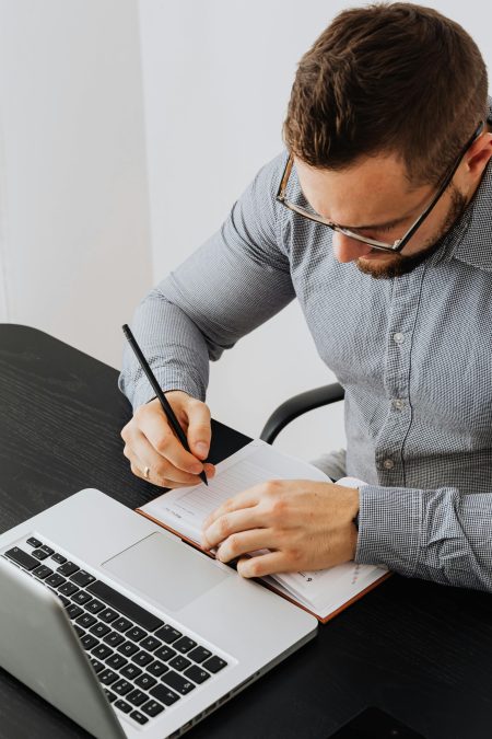 Businessman writing notes on a notebook in a modern office setting with laptop on desk.
