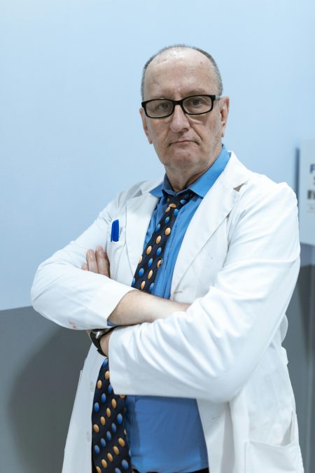 Middle-aged male doctor in white coat, wearing glasses, standing confidently with arms crossed indoors.