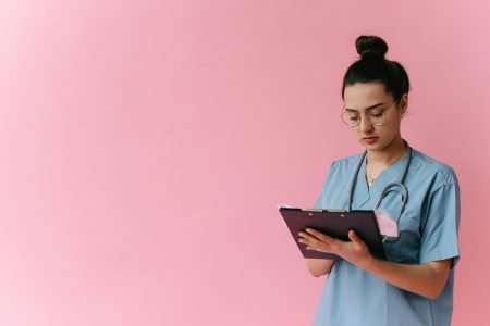 Professional nurse reviewing patient notes on clipboard with pink background, in studio setup.