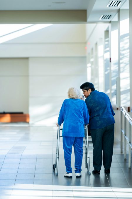 A nurse assists an elderly woman with a walker in a bright hospital corridor.