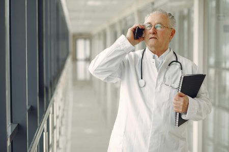 Consultant Occupational Health Physician in Blackpool (Visa Sponsorship Available) Serious mature male doctor in medical uniform standing with stethoscope and notebook while talking on smartphone in clinic corridor