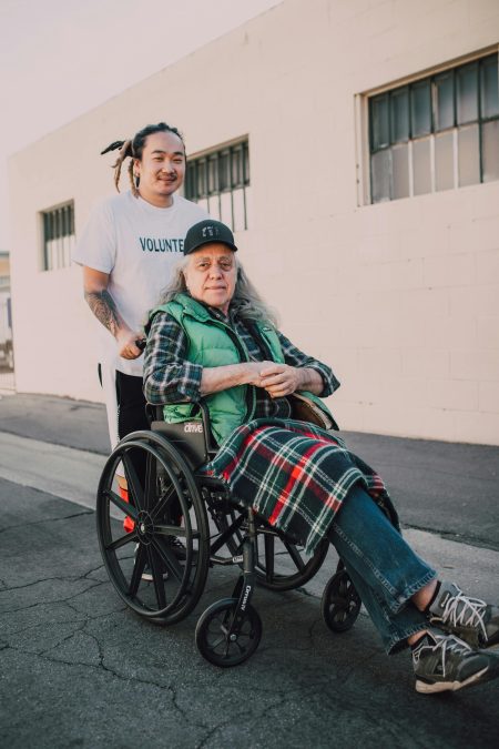 Senior Healthcare Support Worker needed in Bristol (Visa Sponsorship Available) A young volunteer helps an elderly man in a wheelchair on a sunny street.