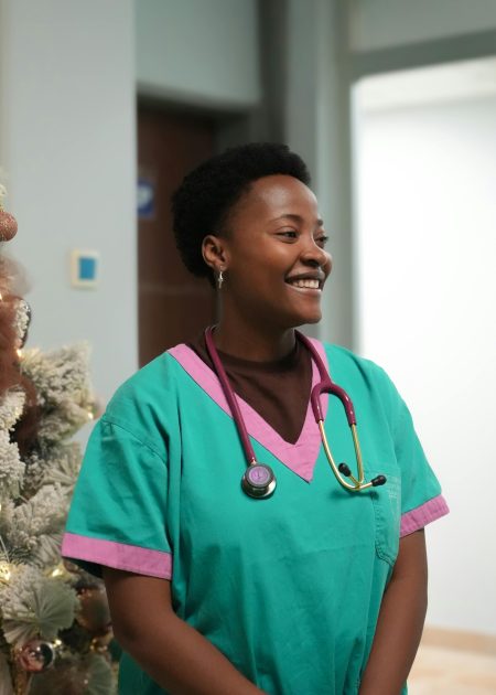 A cheerful nurse stands in a hospital hallway, wearing green scrubs and a stethoscope.