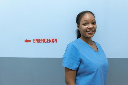 Happy healthcare worker in scrubs standing near emergency room sign.