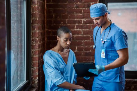 Nurse needed at Bangor (Visa Sponsorship Available) Thoughtful doctor in uniform and gloves showing diagnosis to African American female patient with short hair in blue medical robe in hallway of hospital in daytime