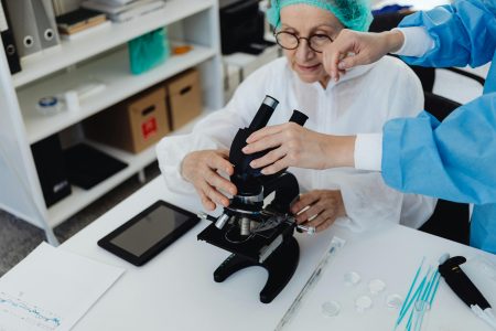 Senior Healthcare Scientist Assistant needed in Plymouth (Visa Sponsorship Available) Senior scientist using a microscope with assistance in a laboratory setting, demonstrating research and analysis.