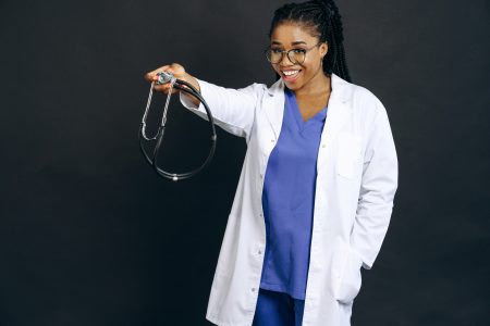 Confident female doctor in white coat smiling and holding a stethoscope against a black background.