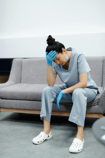 Mental Health Practitioner needed in Redhill (Visa Sponsorship Available) Tired nurse in scrubs sitting on a sofa, looking exhausted with her hand on her head.