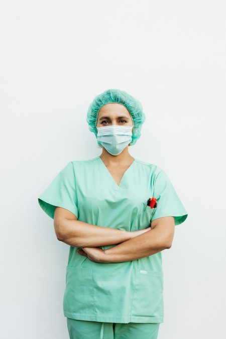 A healthcare professional standing with arms crossed in green scrubs and a protective facemask.