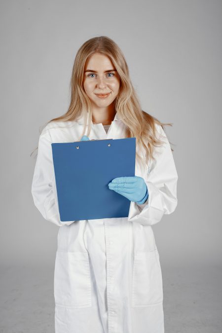 A smiling nurse in a white lab coat and blue gloves holds a clipboard against a neutral background.