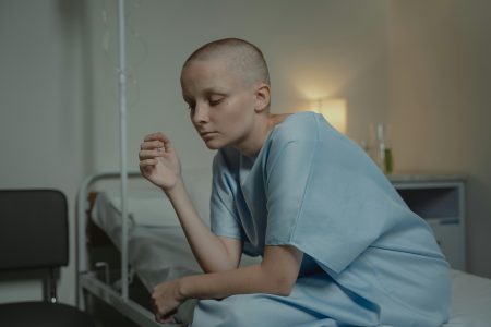 Bald woman sitting in a hospital gown, depicting sadness and contemplation.