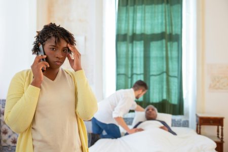 A woman on the phone looks worried as a caregiver assists an elderly person in bed.