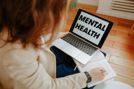 A woman participating in an online therapy session, taking notes with a laptop displaying 'Mental Health'.