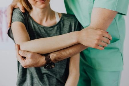 Crop anonymous chiropractor in uniform and wristwatch examining shoulder of smiling faceless woman in casual clothes in doctor office in clinic