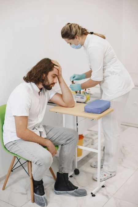A nurse in medical gloves helps a seated patient in a clinic setting.