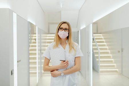 A woman healthcare worker wearing a mask and holding documents in a modern clinic hallway.